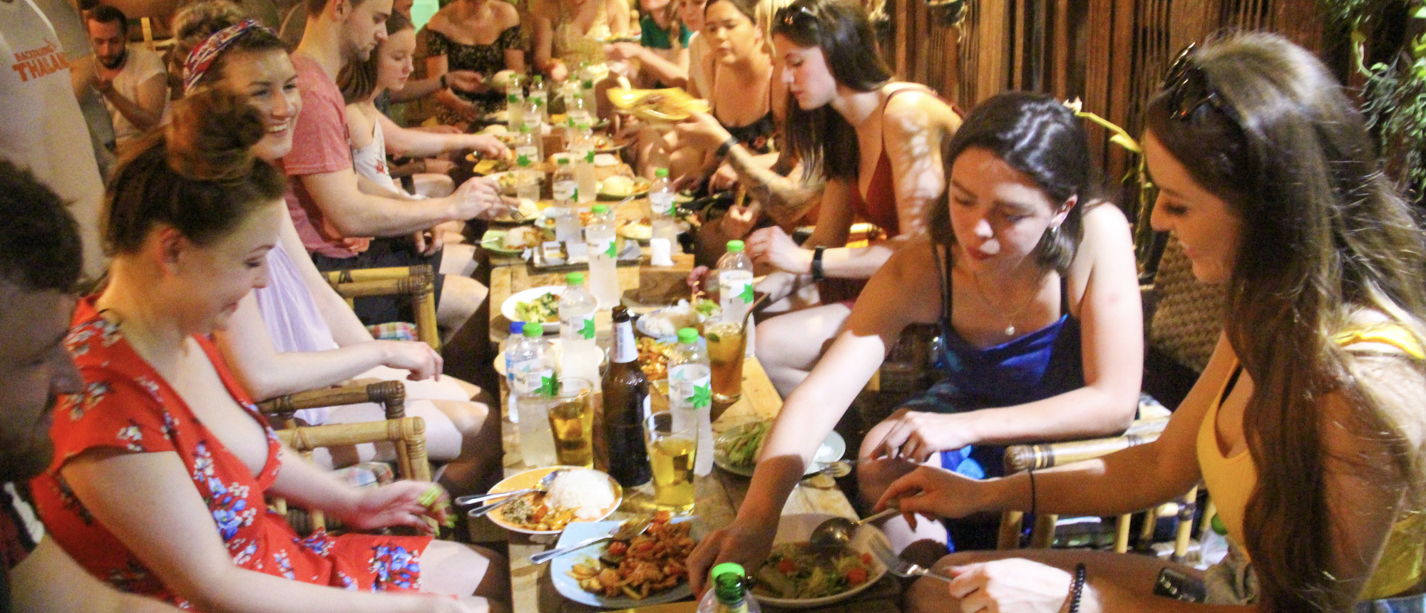 A group of young backpackers enjoying a traditional Thai-style welcome dinner at a long table in Bangkok.