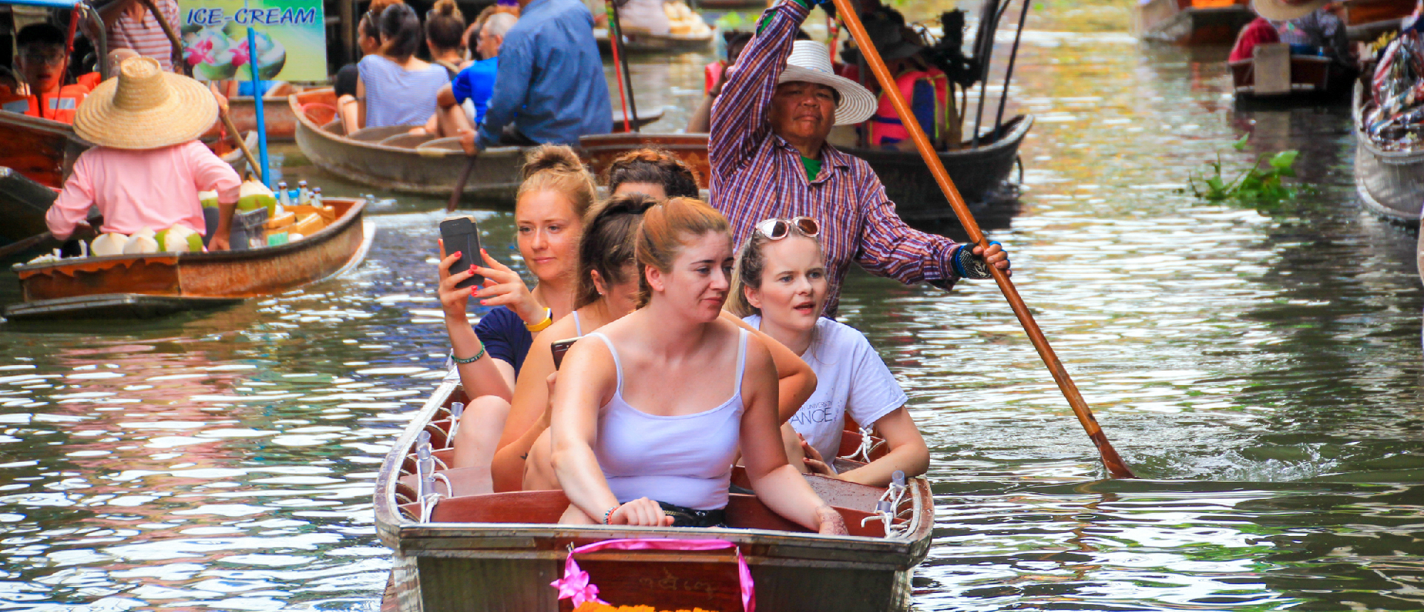 Young women in a boat taking a selfie at the vibrant Amphawa Floating Market in Thailand.