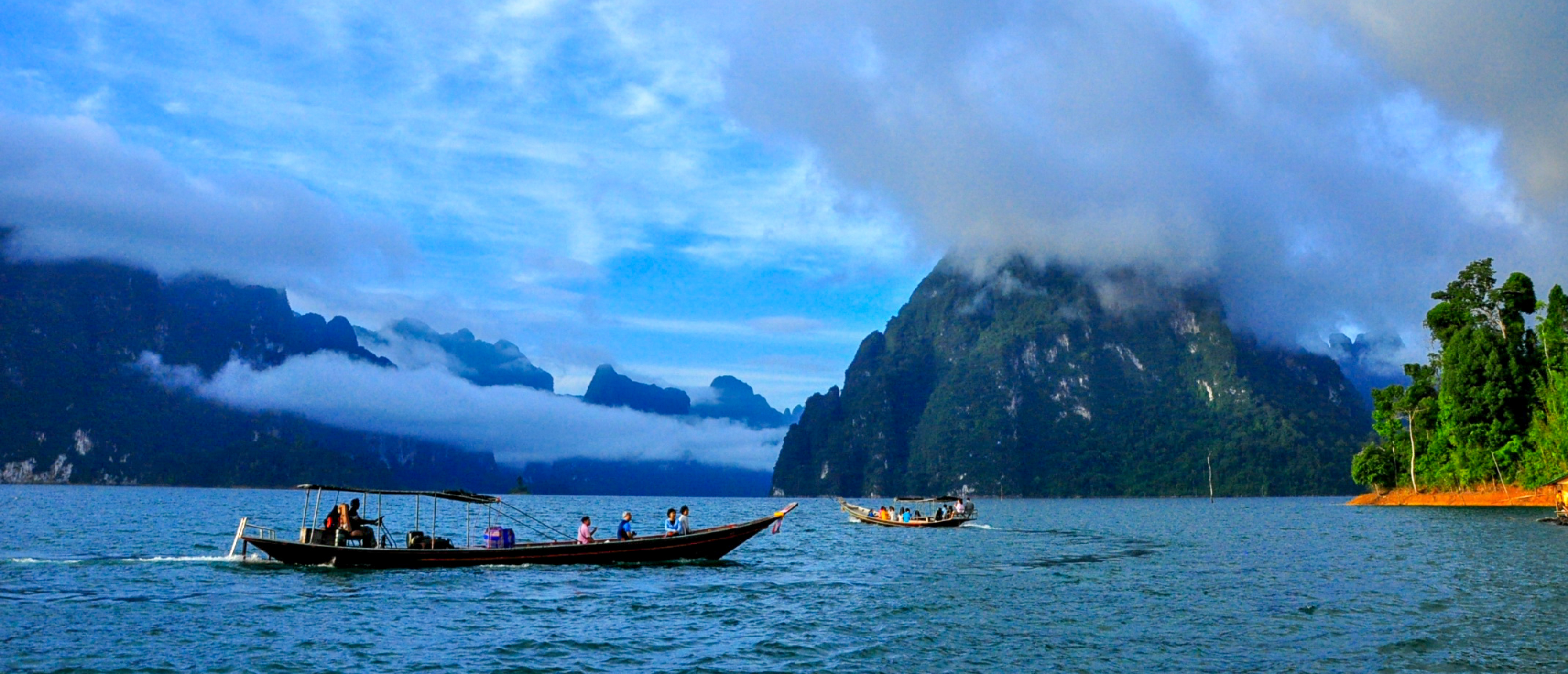 Tourists on longtail boats traverse Cheow Lan Lake, surrounded by misty limestone mountains in Khao Sok.