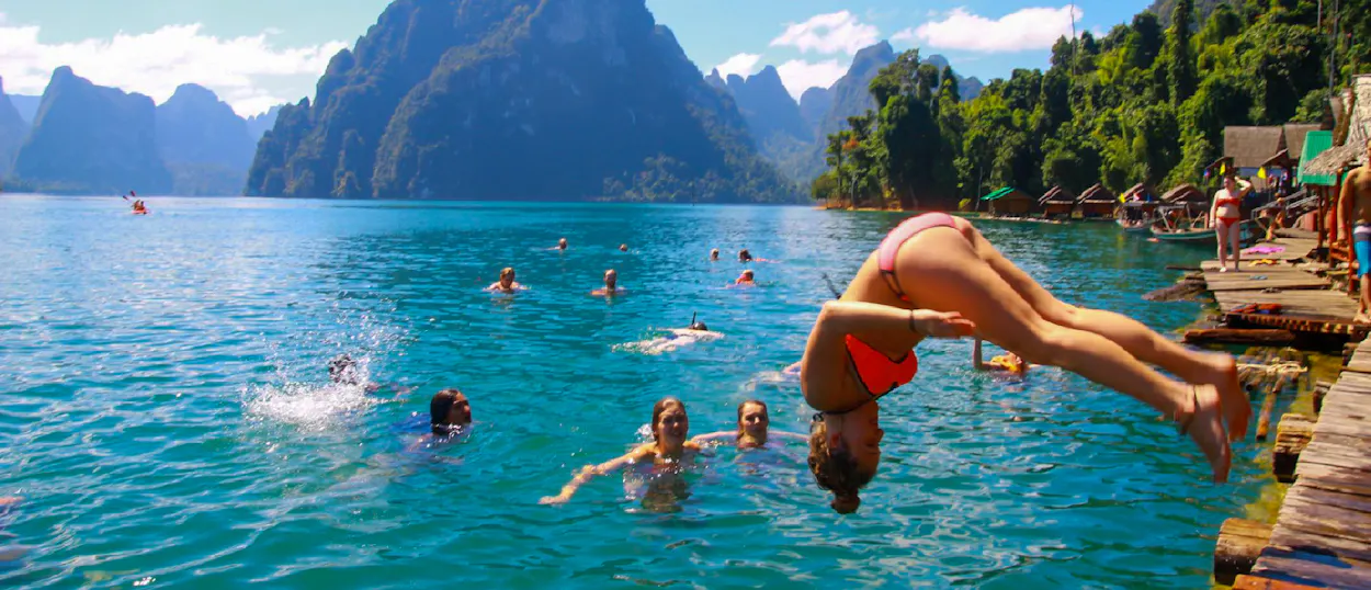 A woman dives headfirst into clear blue water, surrounded by swimmers and limestone mountains in Khao Sok.