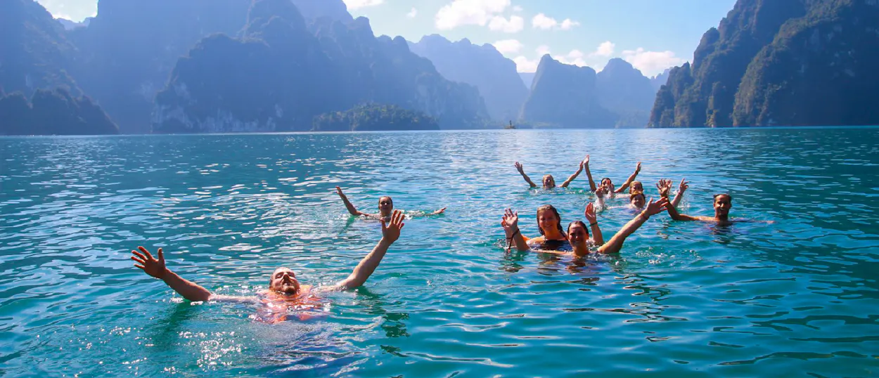Happy backpackers swim in the clear blue waters of Khao Sok Lake, surrounded by towering limestone mountains.