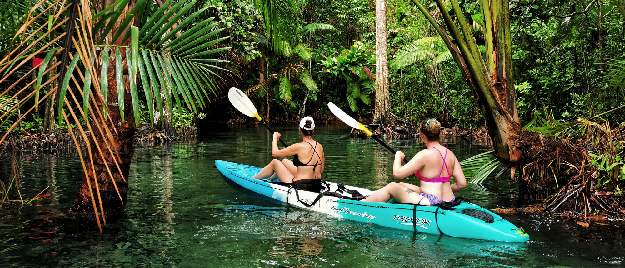 Two women paddle a kayak through a clear emerald river surrounded by lush jungle in Krabi.