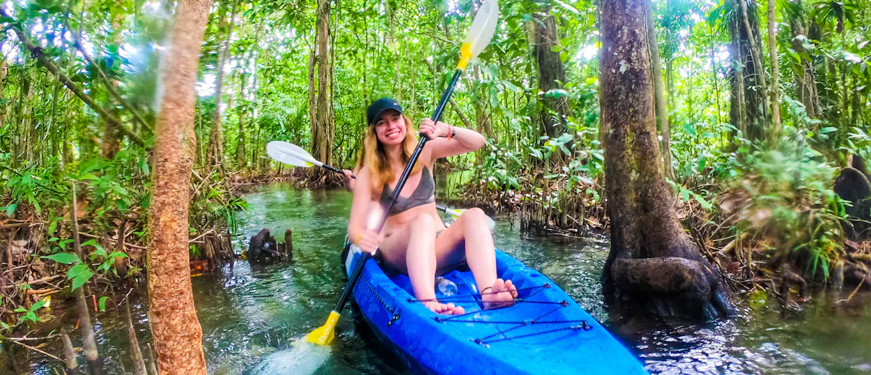A smiling woman paddles a blue kayak through a clear emerald river in Krabi's mangrove forest.