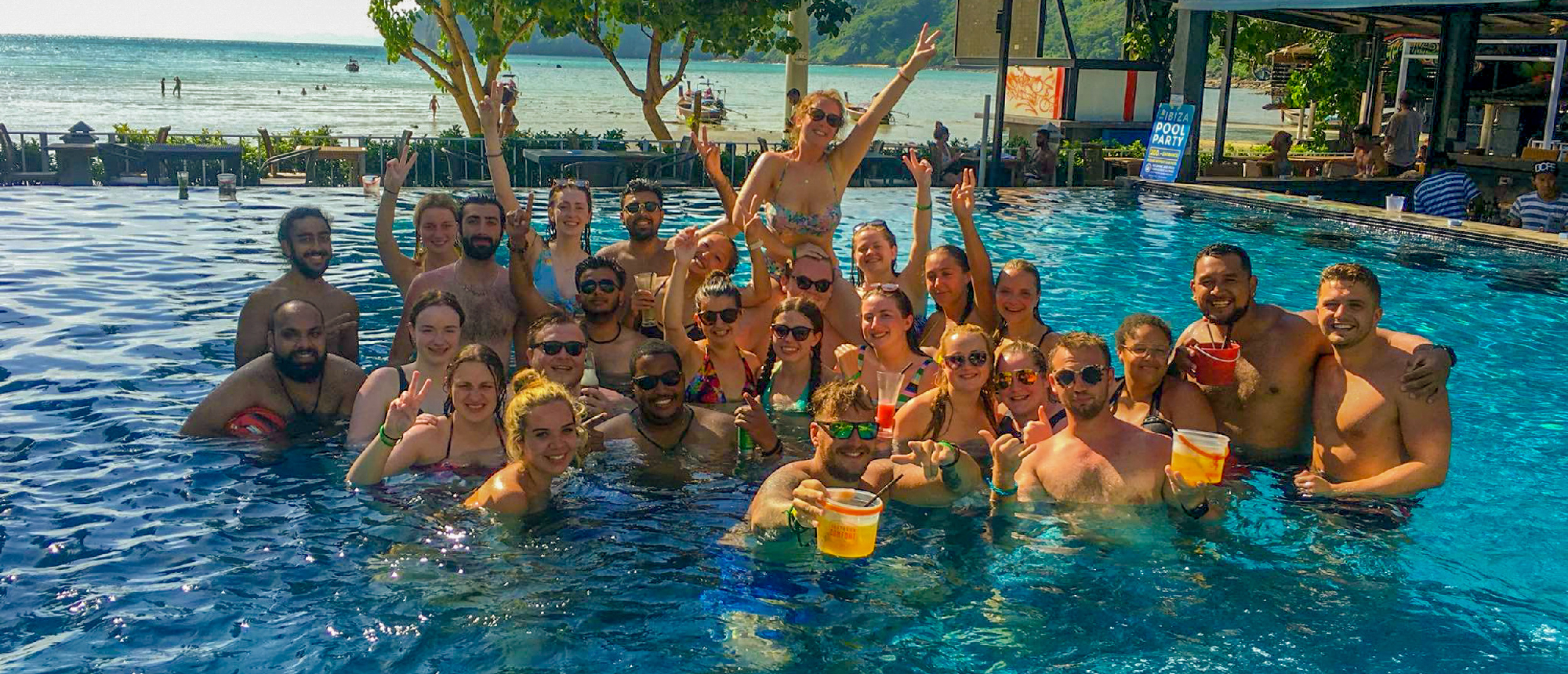 A large group of happy backpackers enjoying a lively pool party with drinks in Phuket, Thailand.