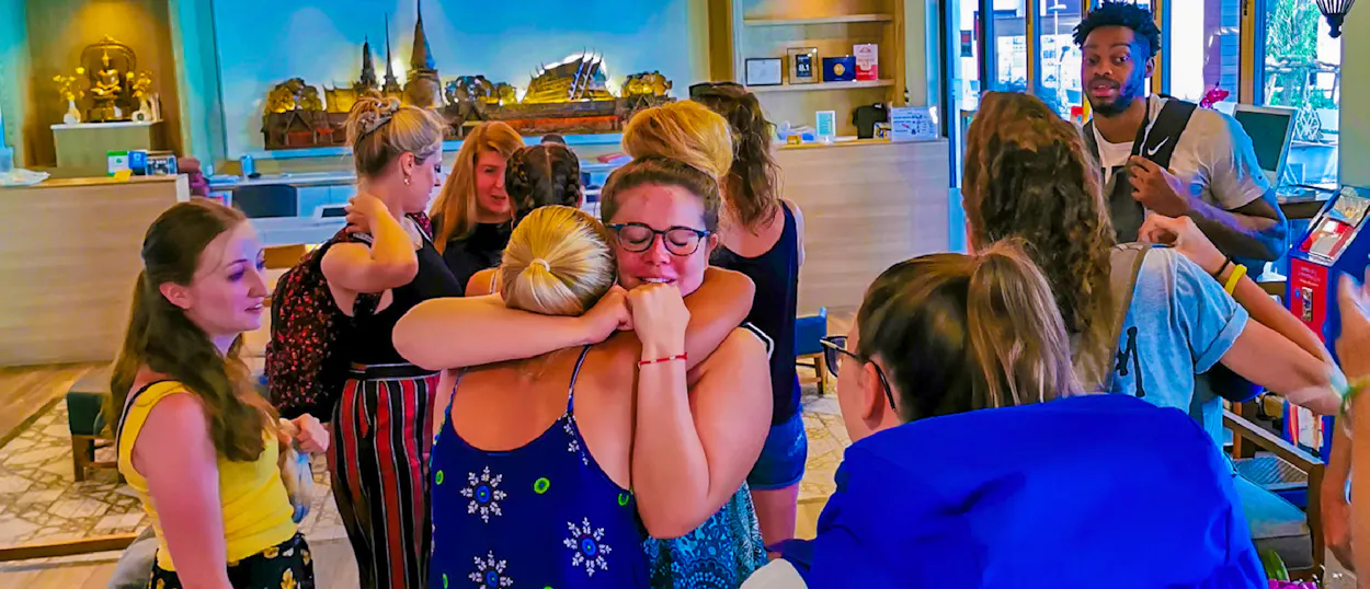 Two women share an emotional hug, surrounded by their travel group in a Phuket hotel.