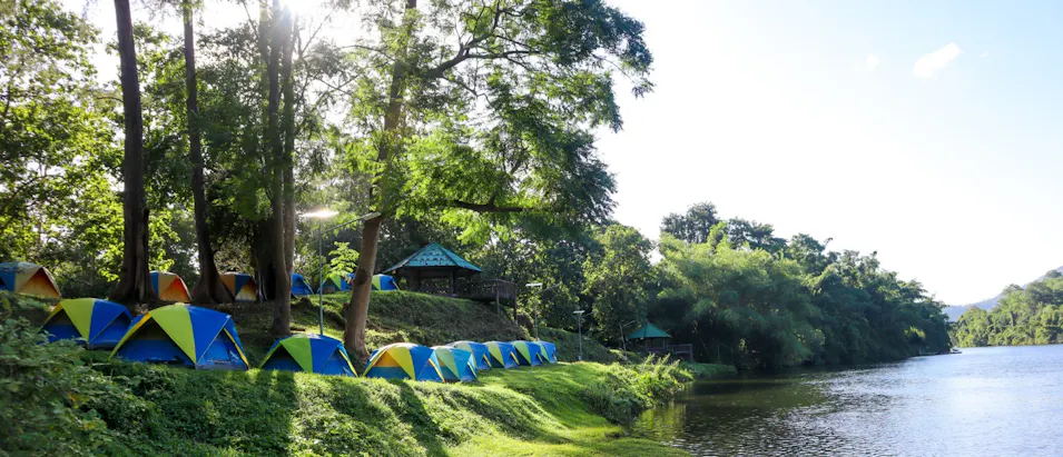 Many blue and green tents line a lush bank beside a calm lake in Thailand.