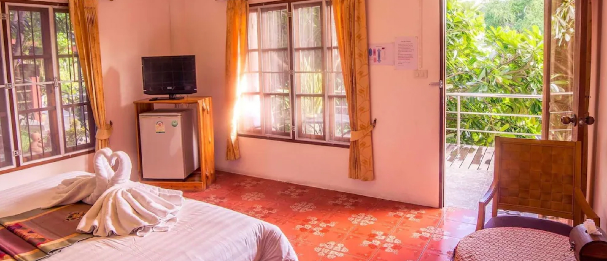 A clean hotel room in Thailand with a bed, TV, and open door to a green balcony.