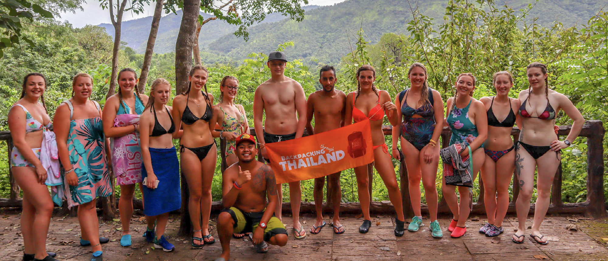 A group of happy backpackers in swimwear pose with a flag in Thailand's Erawan National Park.