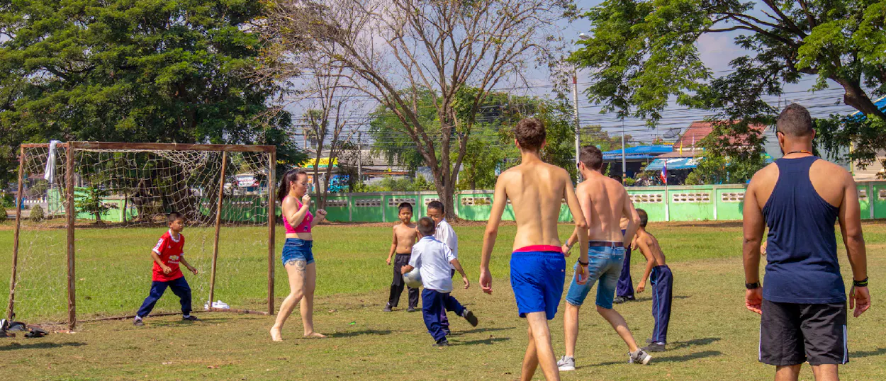Backpackers and local Thai children happily play football together on a sunny school field in Amaphawa.