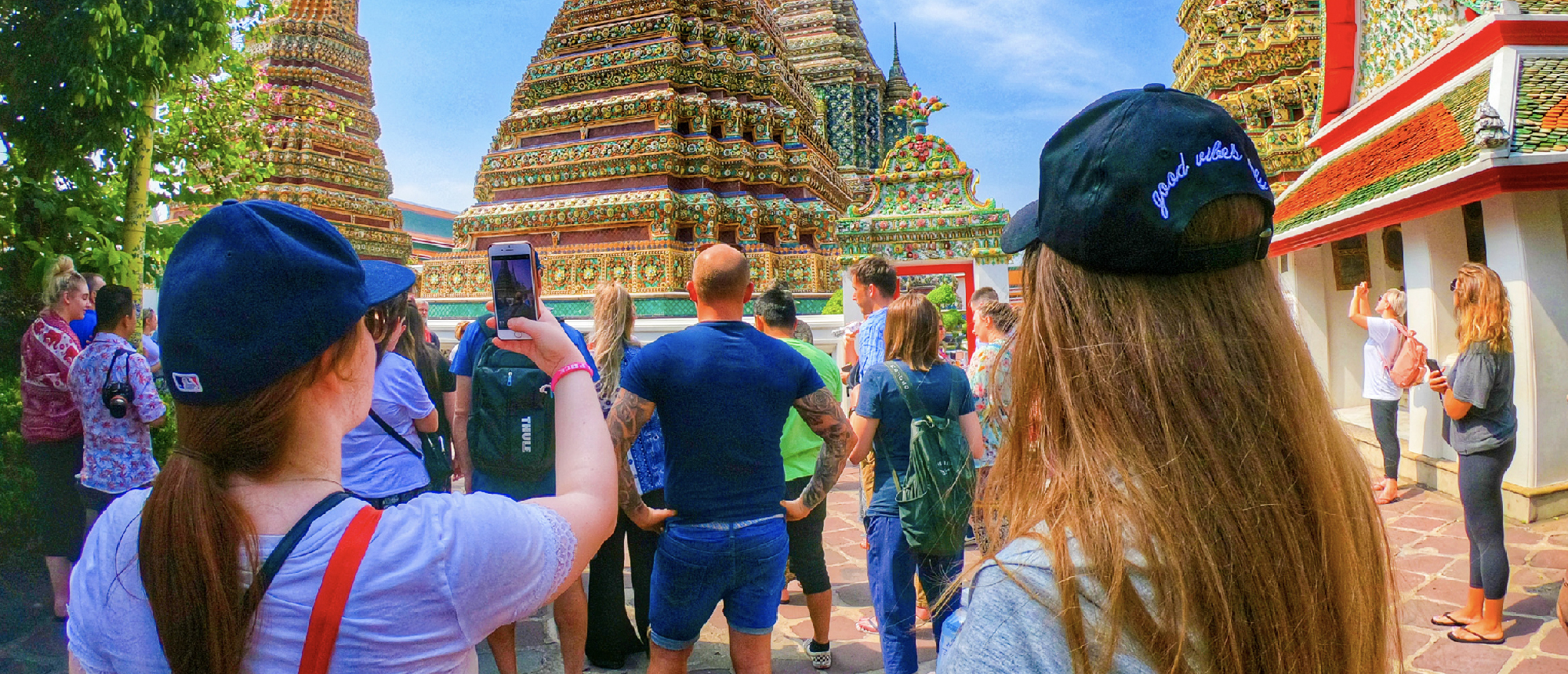 A group of tourists explores and photographs ornate temple spires under a bright blue sky in Bangkok.