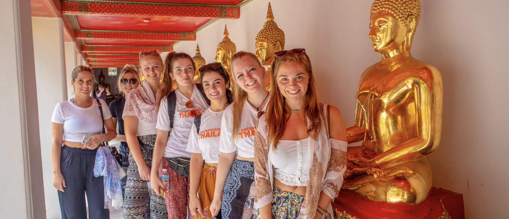A group of smiling young women posing in a temple with golden Buddha statues in Bangkok.