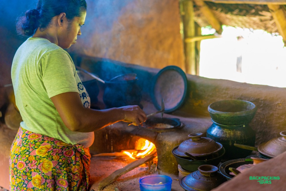 A local woman cooking traditional food over a clay oven in a rustic Sri Lankan setting.