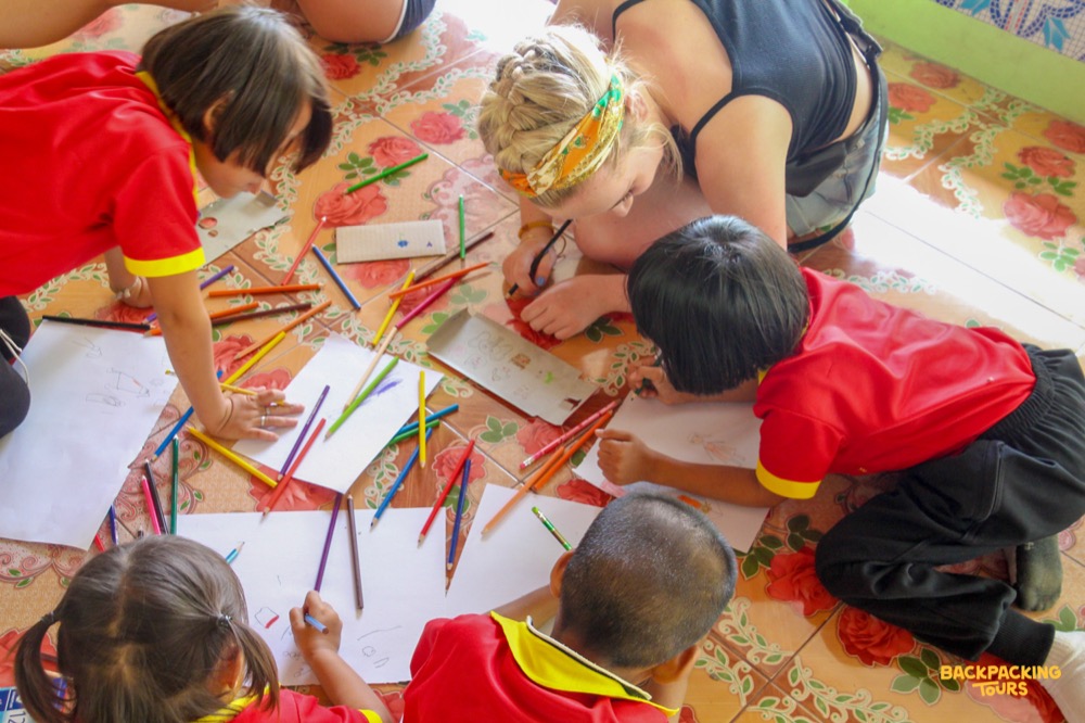 A volunteer and four children drawing with colored pencils on a patterned floor in a Thai school.