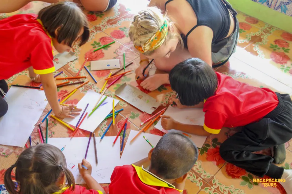 A volunteer and four children drawing with colored pencils on a patterned floor in a Thai school.