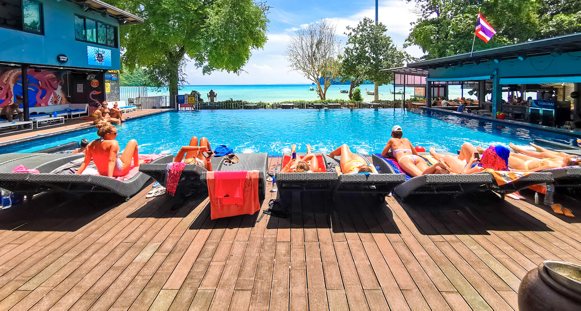 Sunbathers relax by a vibrant blue pool with a clear view of the turquoise ocean in Thailand.