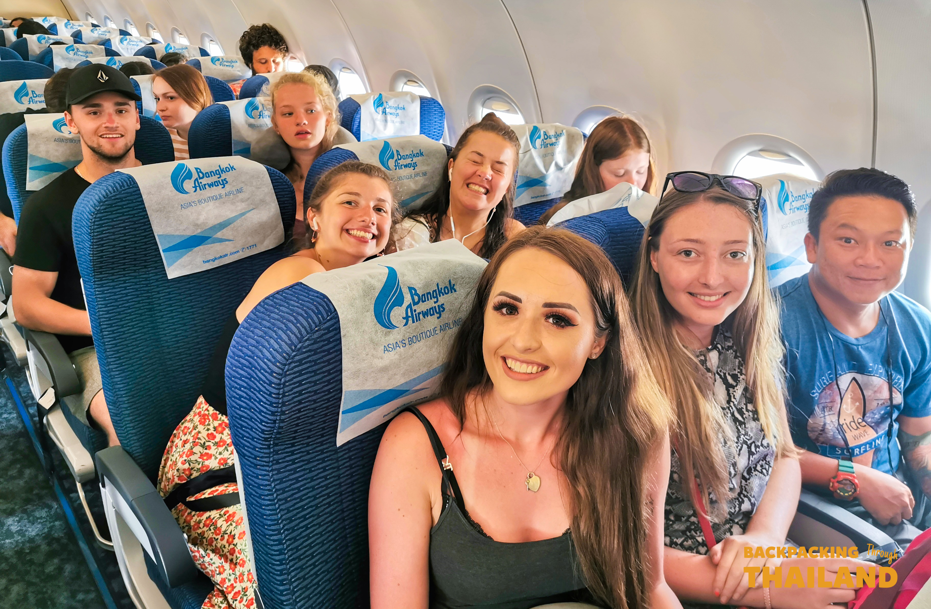 Happy young backpackers smiling and laughing together inside a Bangkok Airways plane cabin.