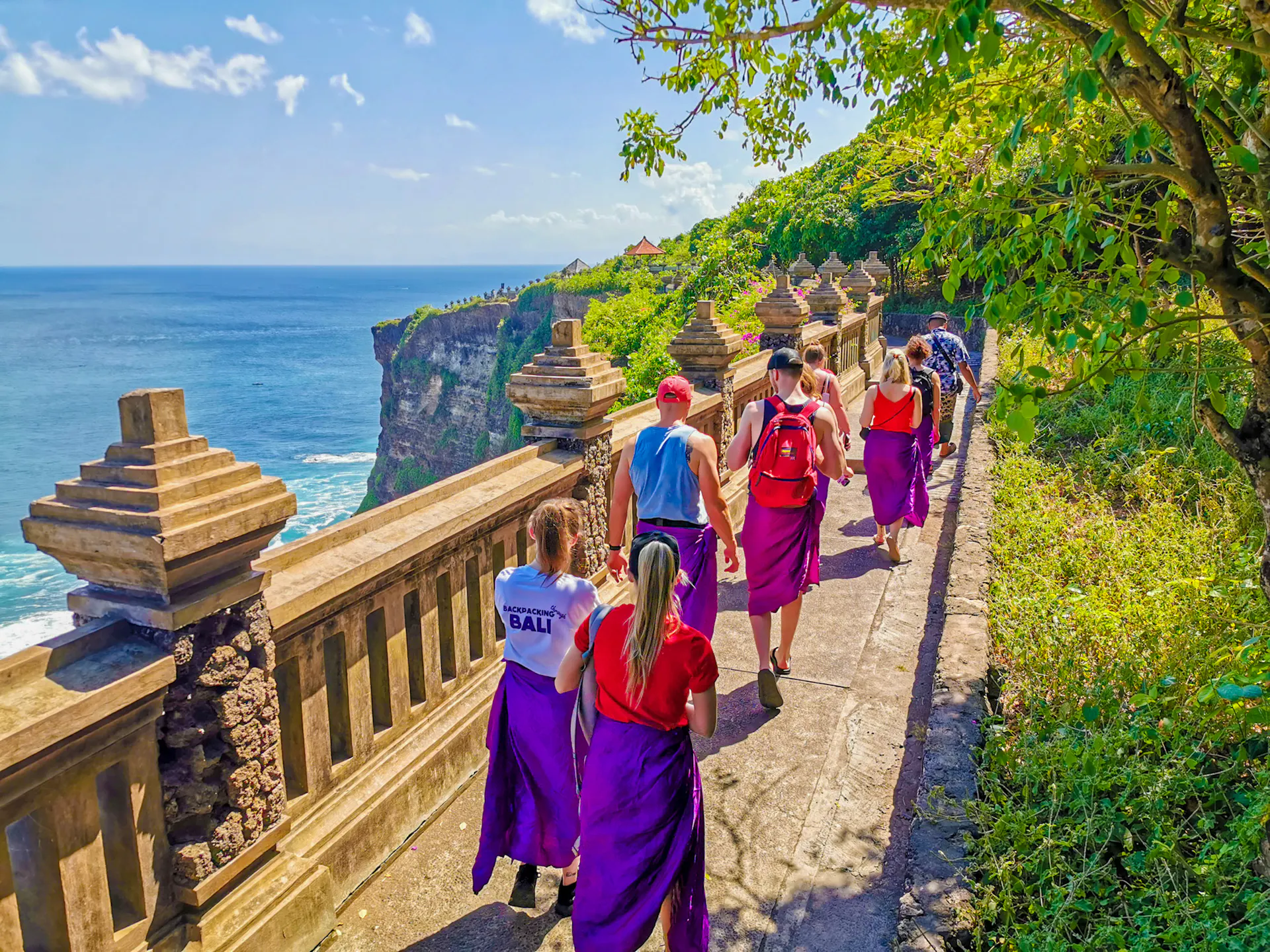 Tourists in purple sarongs walk a scenic cliffside path overlooking the ocean at a Balinese temple.