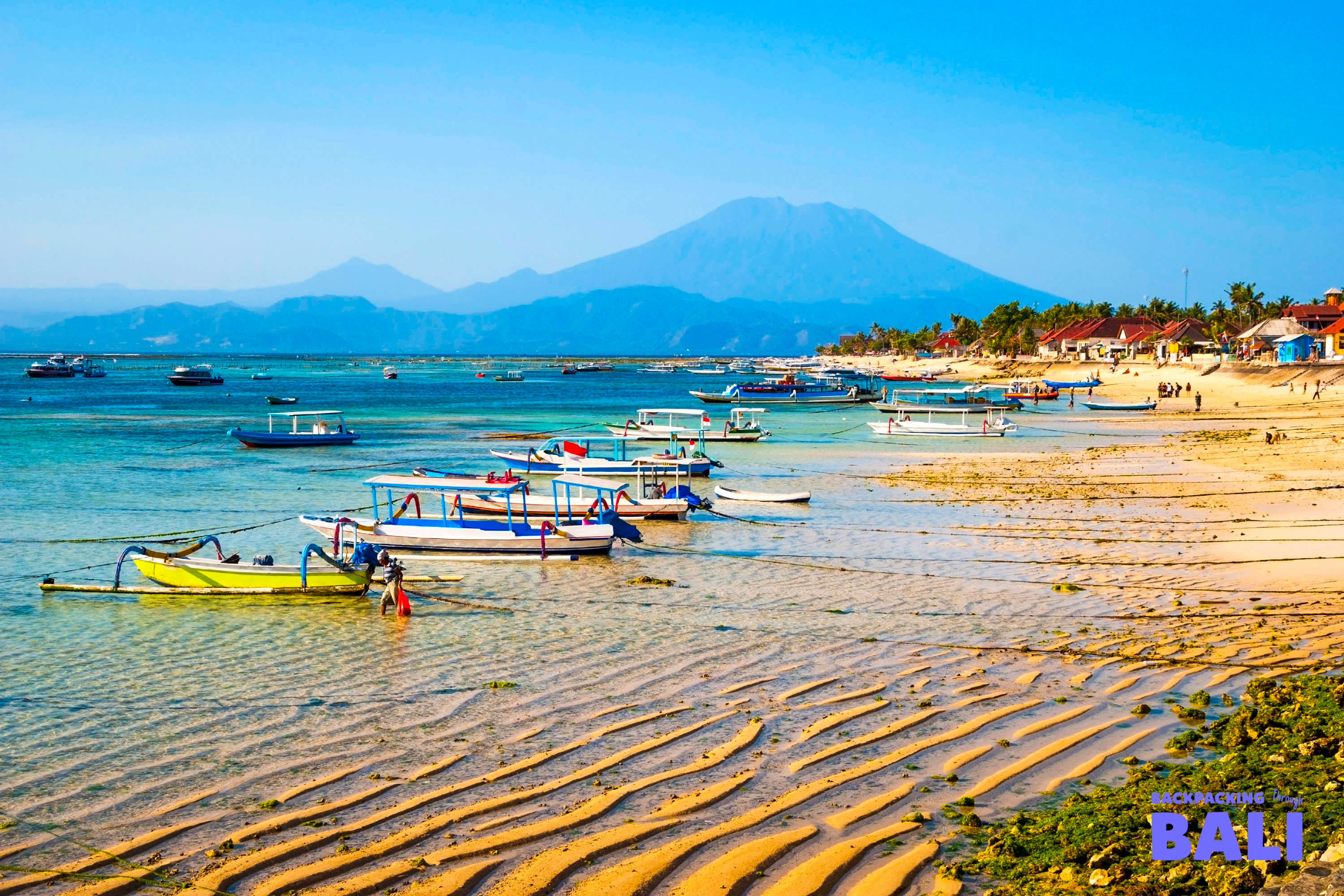 Colorful traditional boats rest on a sandy beach with clear water and a majestic volcano on Nusa Lembongan.