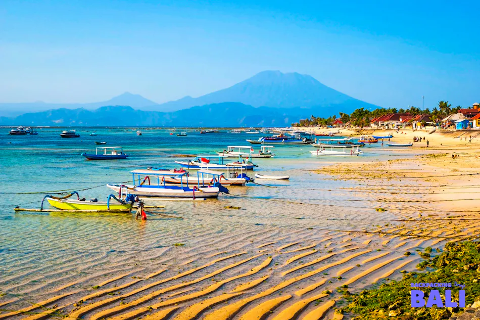 Colorful traditional boats rest on a sandy beach with clear water and a majestic volcano on Nusa Lembongan.