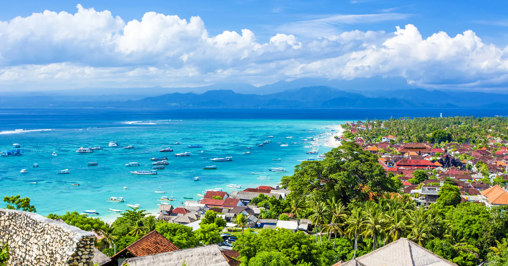 Scenic aerial view of Nusa Lembongan's turquoise bay filled with boats, a coastal village, and distant mountains.