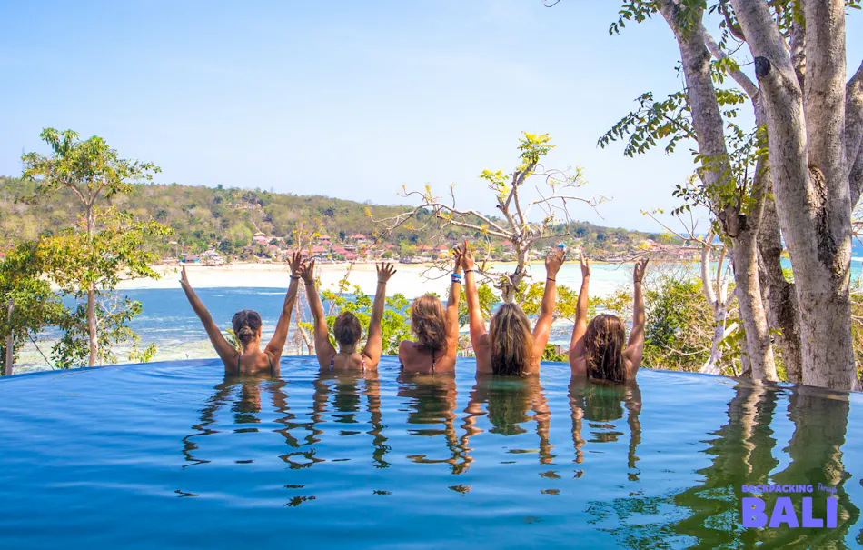 Five friends joyfully raise their arms in an infinity pool overlooking Nusa Lembongan beach.