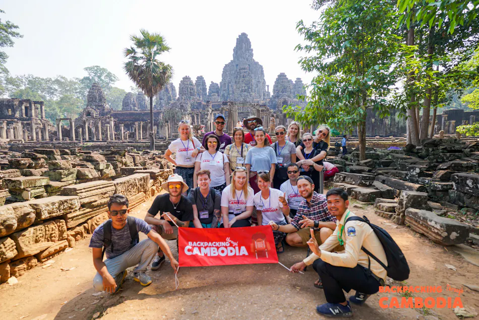 Smiling backpackers proudly hold a 'Backpacking Cambodia' banner before the ancient Bayon temple.