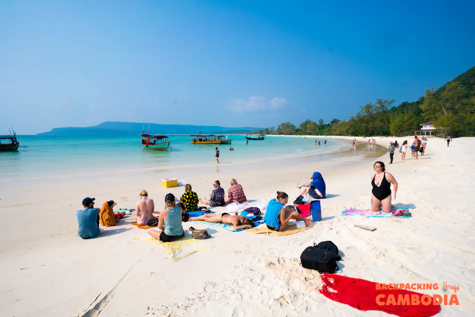 Backpackers sunbathe and swim on a beautiful white sand beach with clear turquoise water in Cambodia.