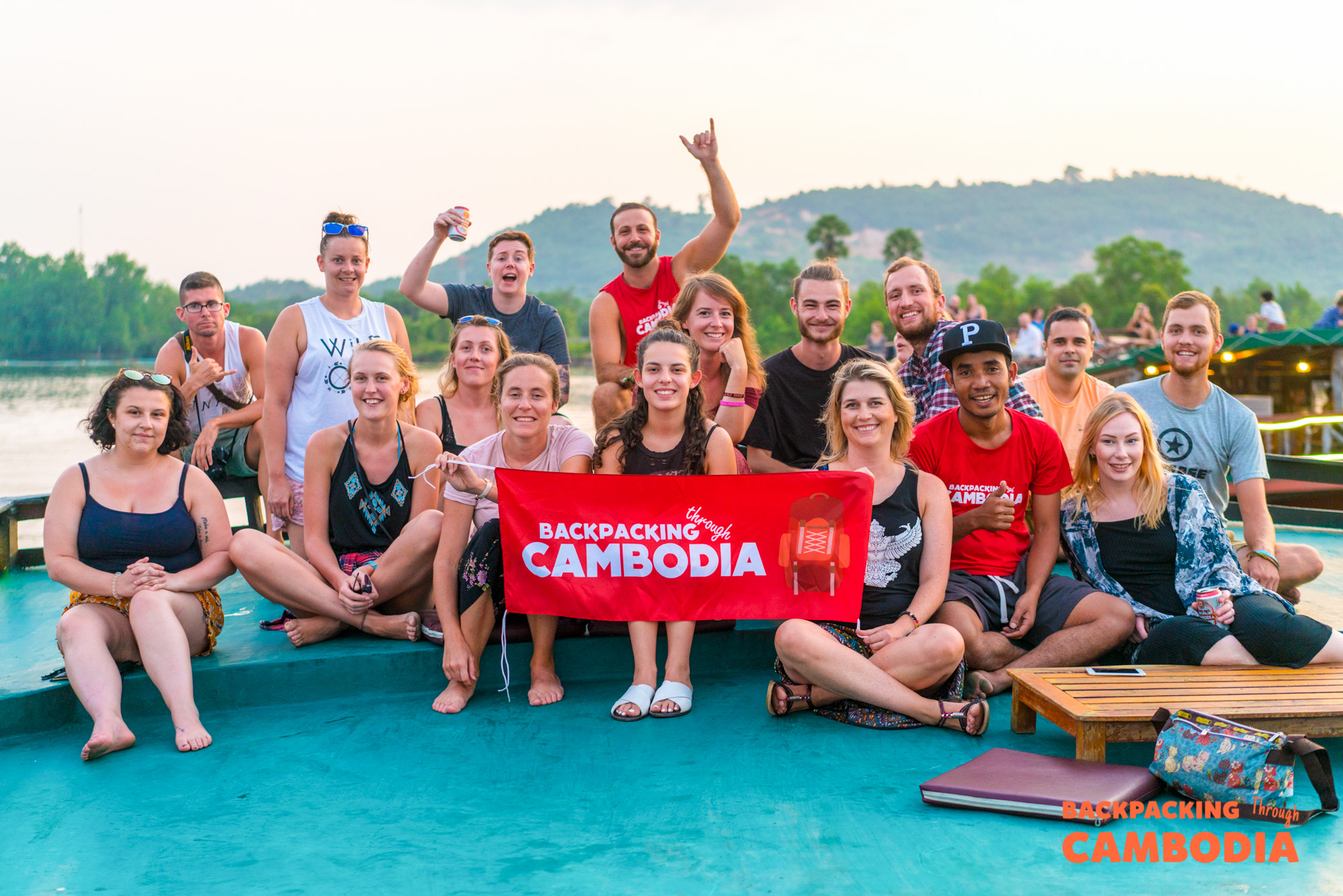 A large group of happy backpackers holding a "Backpacking Cambodia" banner on a boat at sunset.