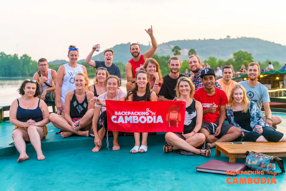 A large group of happy backpackers holding a "Backpacking Cambodia" banner on a boat at sunset.