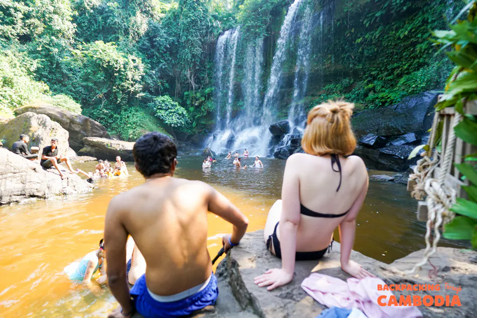 Backpackers sit on rocks, enjoying a refreshing tropical waterfall and swimming hole in Cambodia.