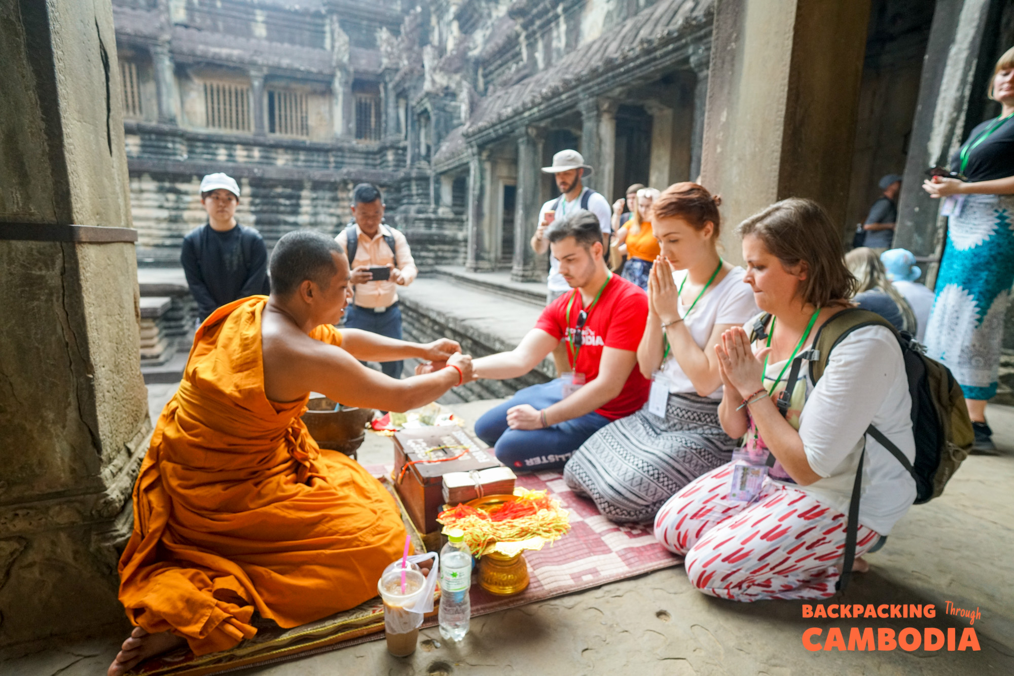 A Buddhist monk blesses kneeling tourists at Angkor Wat temple in Cambodia, surrounded by offerings.