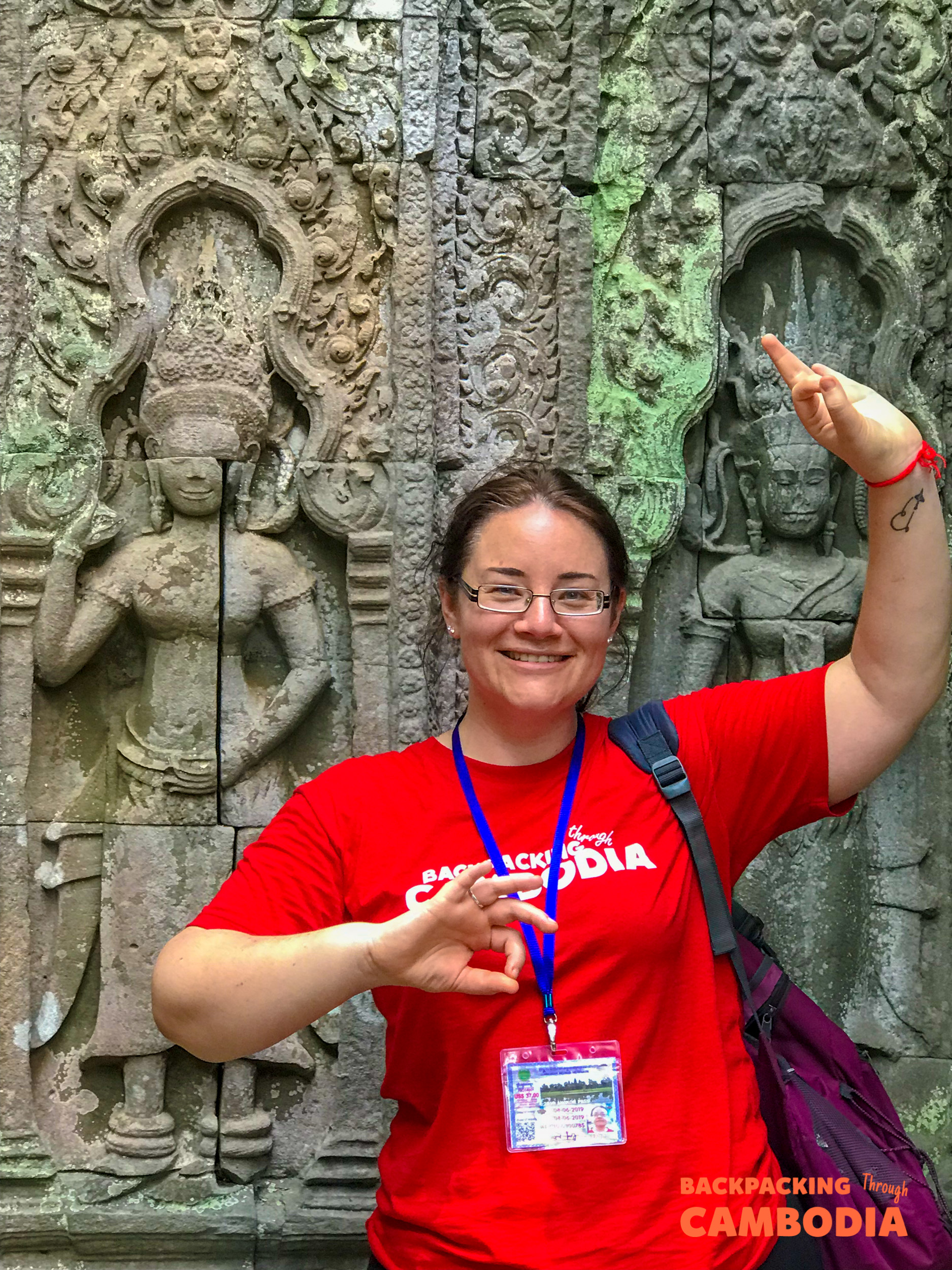 A smiling woman poses in front of ancient moss-covered stone carvings at a Cambodian temple.