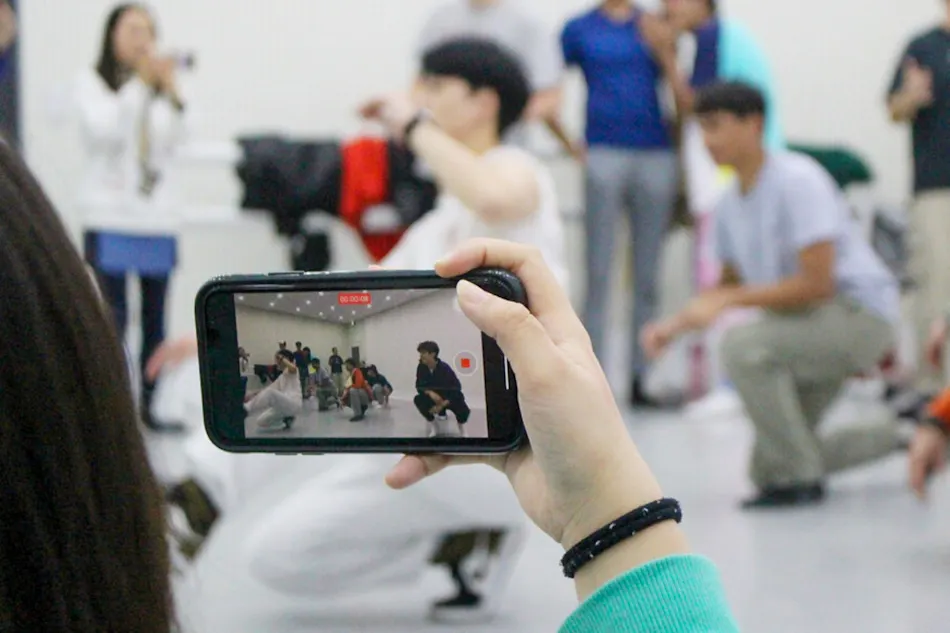 A person records a K-pop dance class with a smartphone, capturing energetic dancers in a studio.