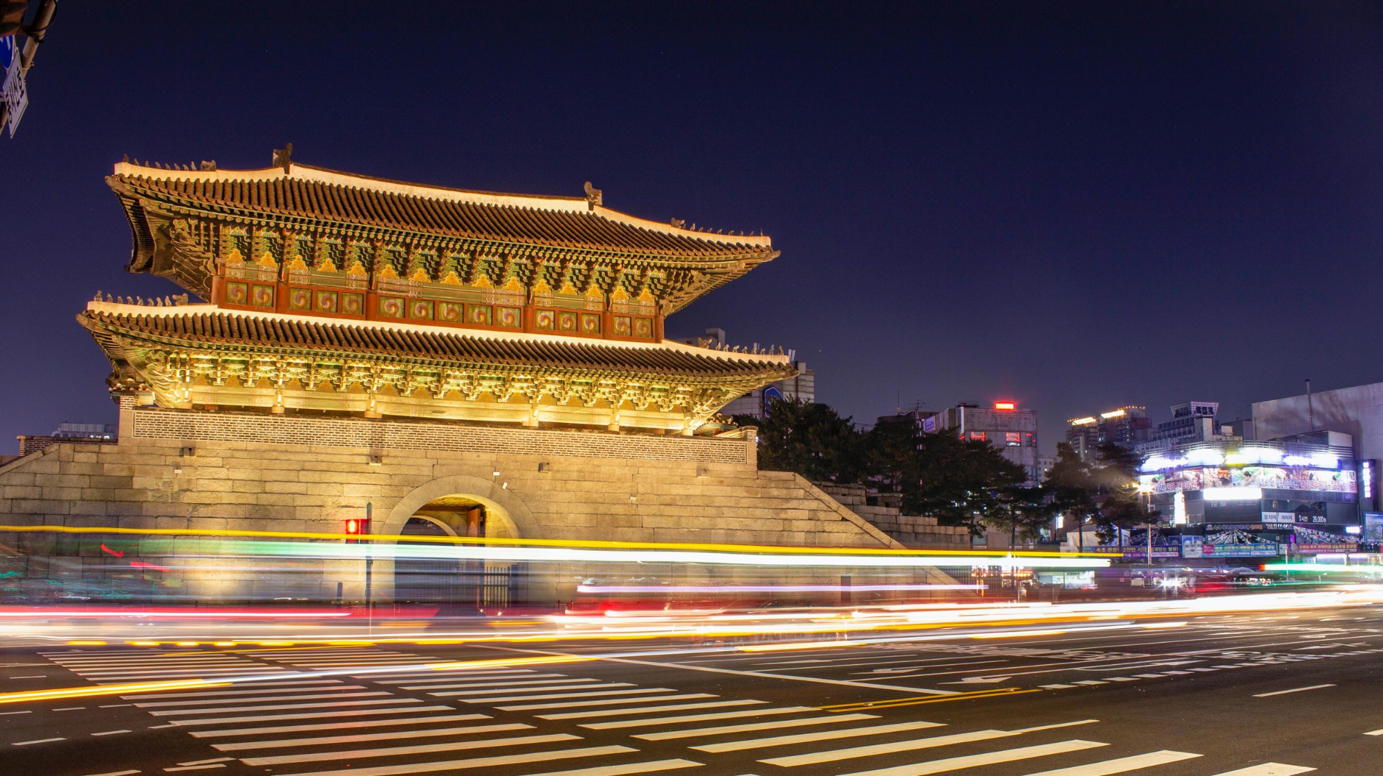 An ornate traditional Korean gate in Seoul illuminated at night with vibrant light trails from traffic.