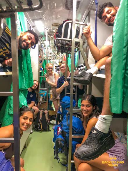 Happy backpackers smile from bunks and seats inside a busy Thai night train carriage.