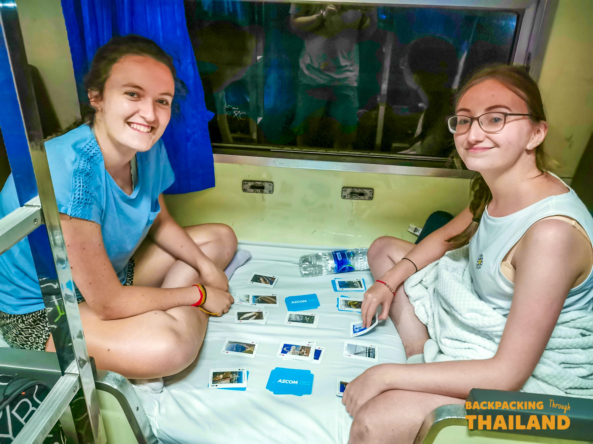 Two smiling women playing cards on a bunk bed inside a night train in Thailand.