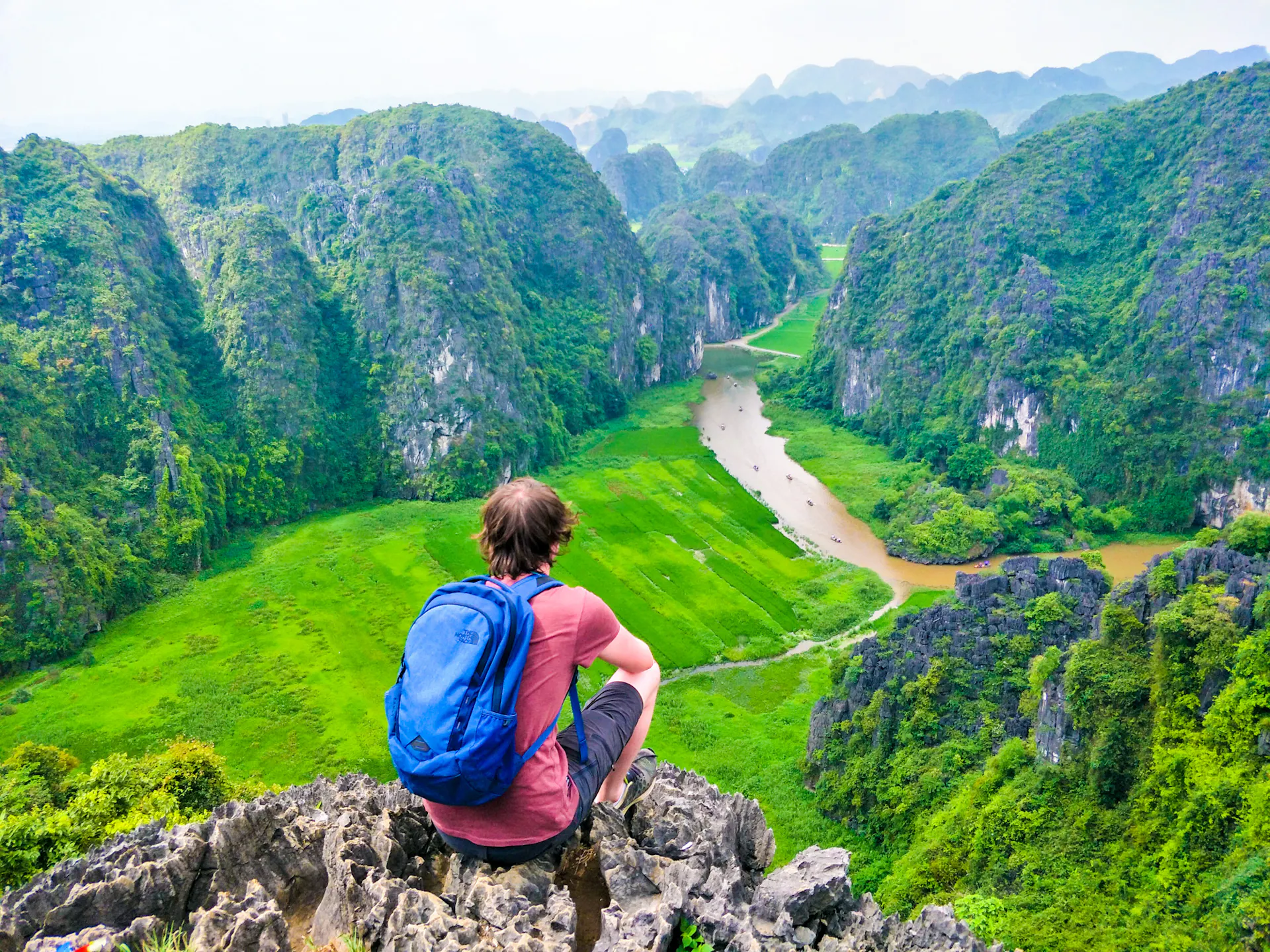 Backpacker sits on a rocky peak, admiring the stunning green karst landscape and winding river in Vietnam.