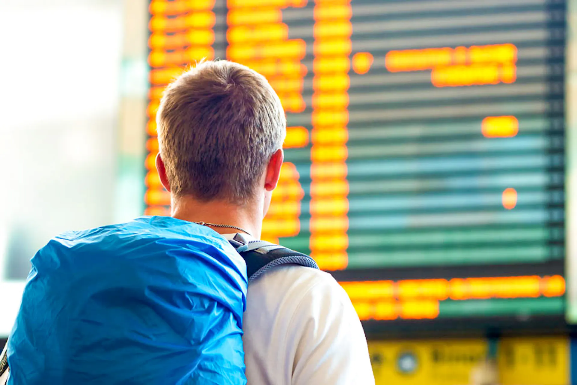 A male traveler with a blue backpack checking flight information on a large airport screen.
