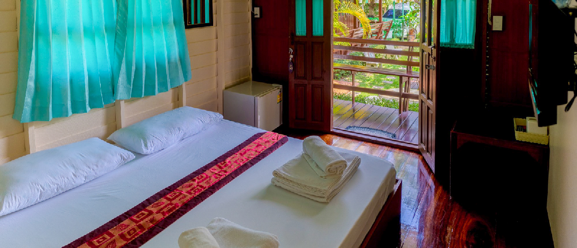 A welcoming bedroom in Thailand with a double bed, patterned runner, and open door to a green balcony.