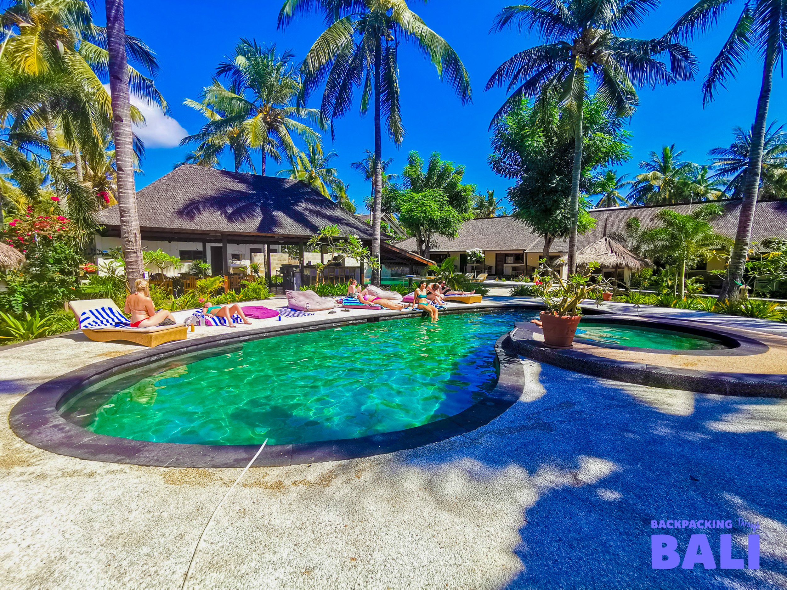 People relax by a vibrant green pool, surrounded by palm trees and bungalows on sunny Gili Trawangan.