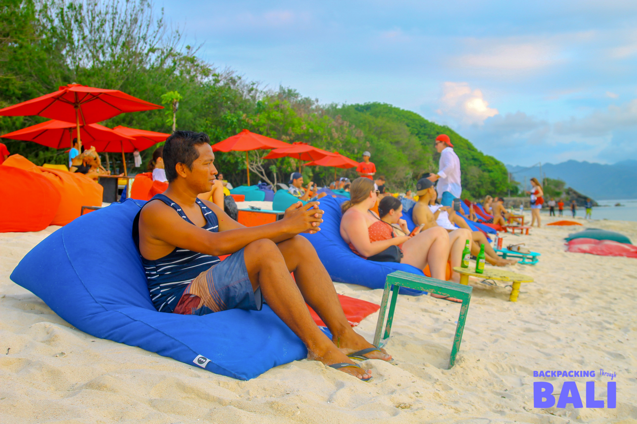 Tourists relax on colorful beanbags on a white sandy beach with red umbrellas on Gili Trawangan.