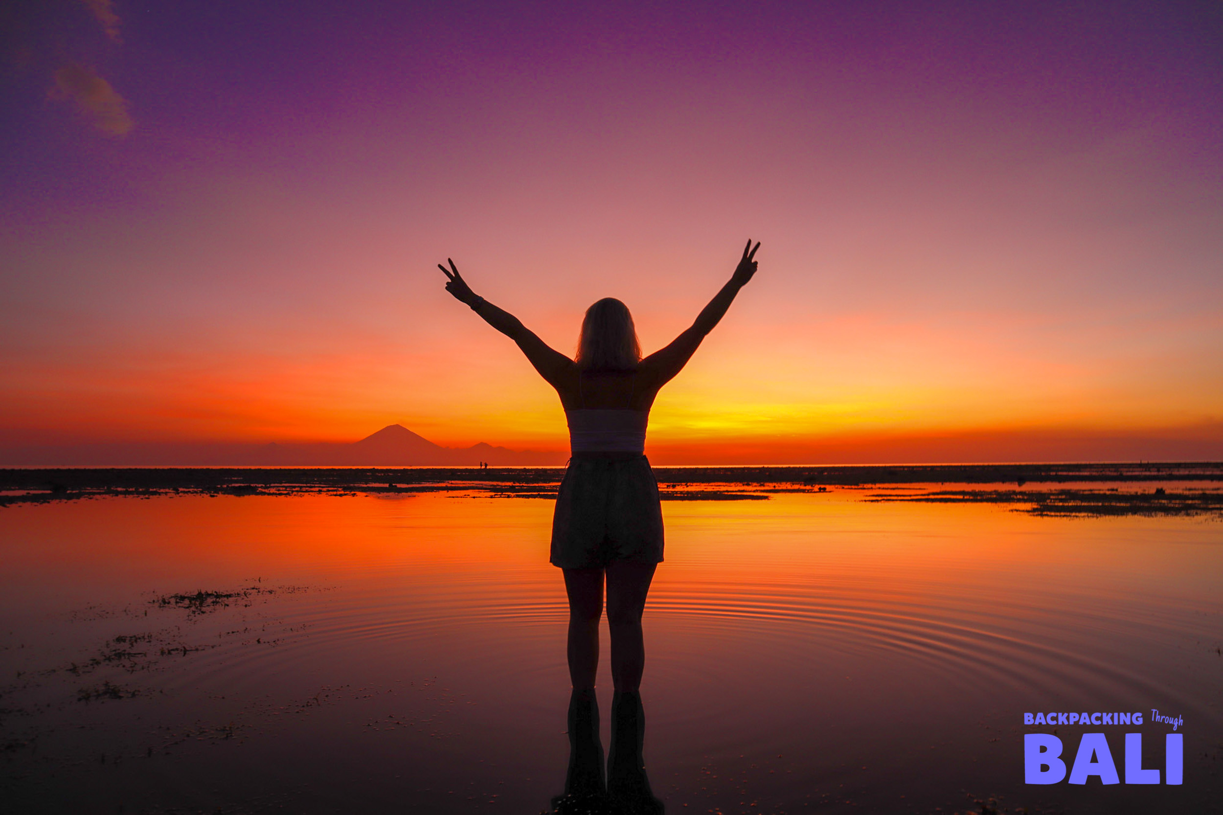 A person celebrates a breathtaking Gili Trawangan sunset with arms raised in the shallow ocean.