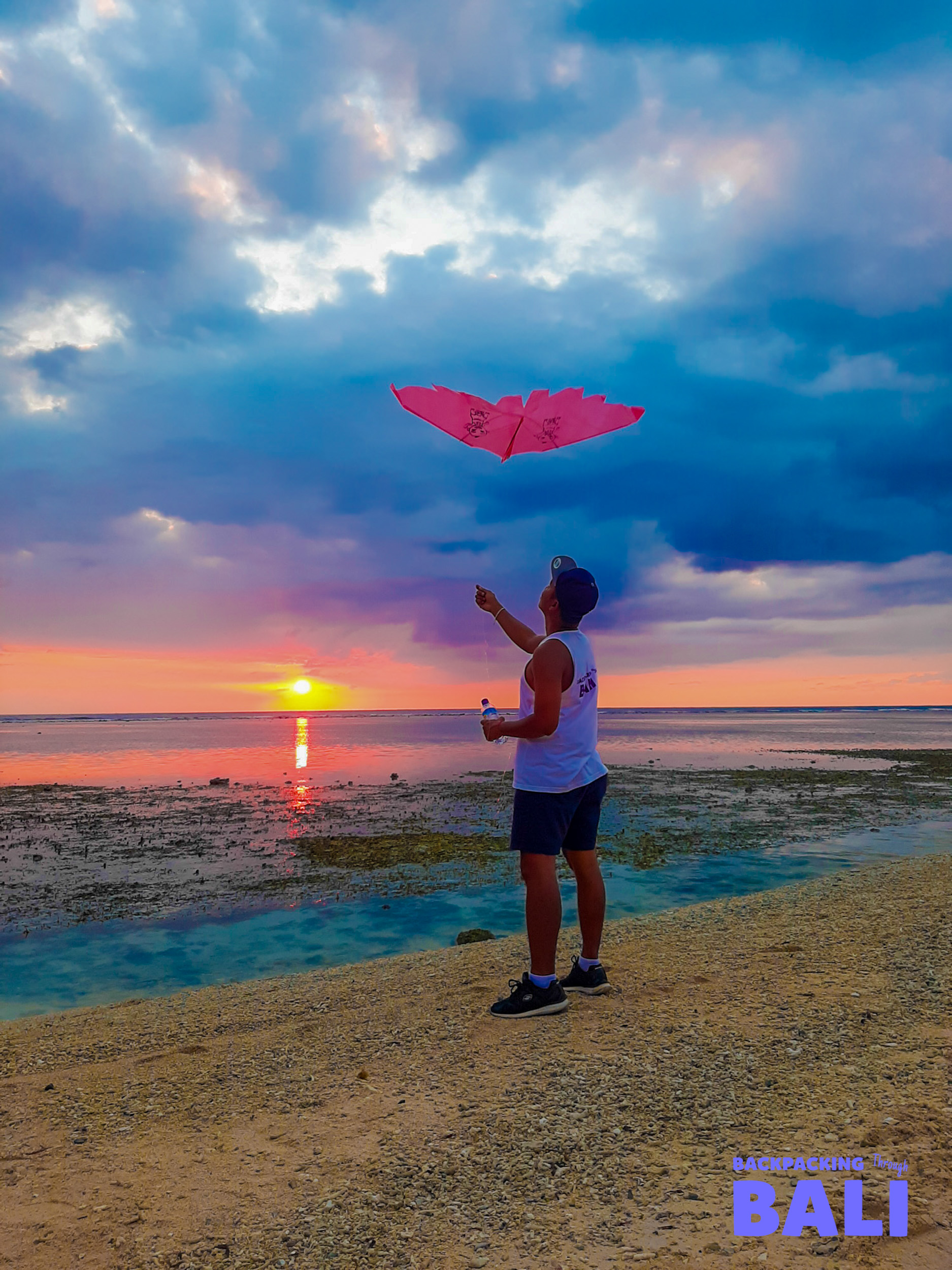 A man flies a pink kite on Gili Trawangan beach at sunset, under a vibrant, cloudy sky.
