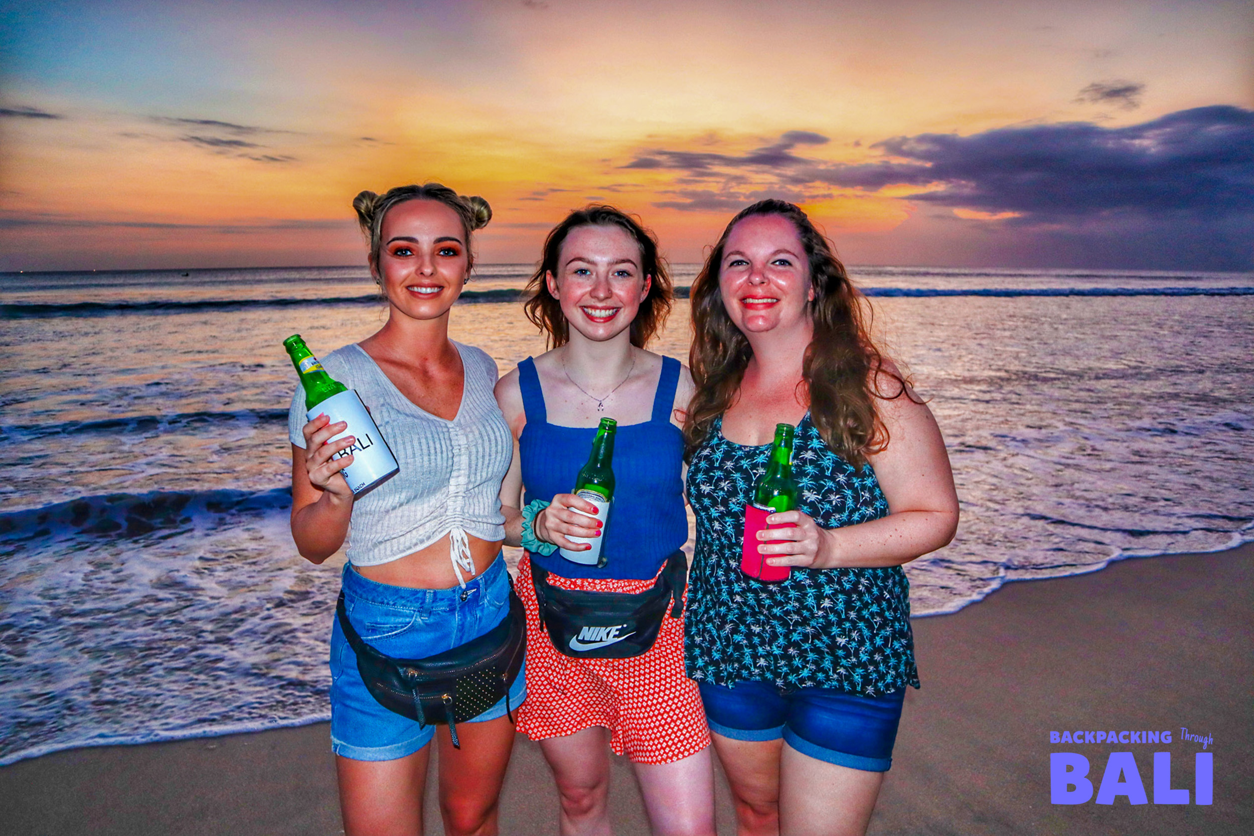 Three friends smile with drinks on Canggu beach, enjoying a beautiful Bali sunset.