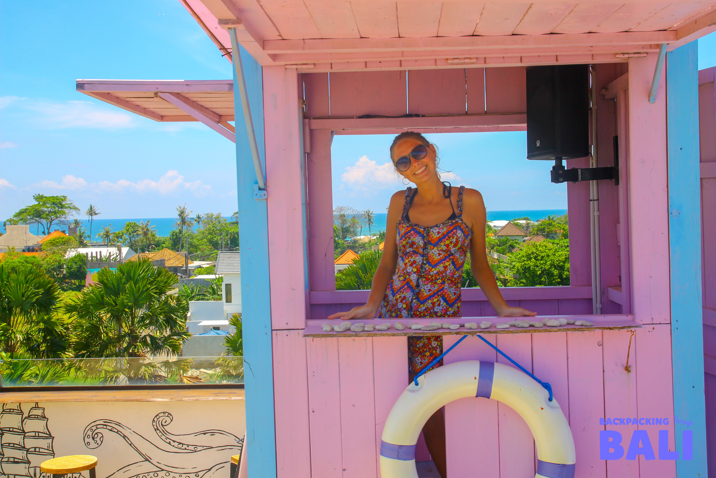 A cheerful woman smiles from a bright pink bar overlooking the beautiful Bali coast.