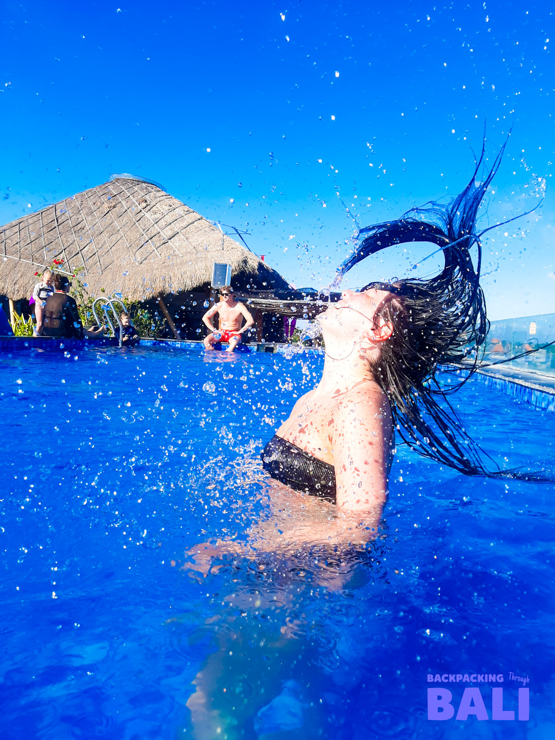 A woman playfully flips her wet hair, splashing water in a bright blue pool in Canggu, Bali.