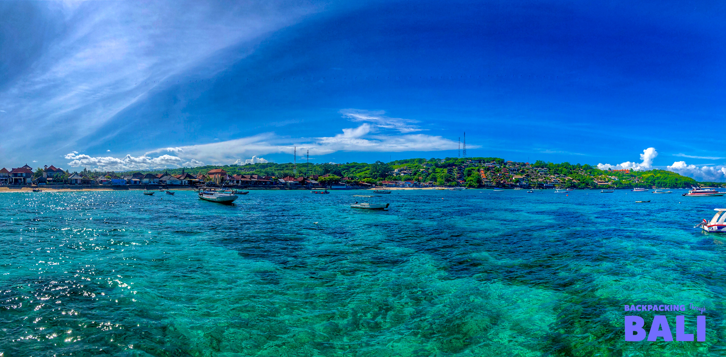 Boats on crystal clear turquoise water with the tropical island coastline of Nusa Lembongan under a blue sky.