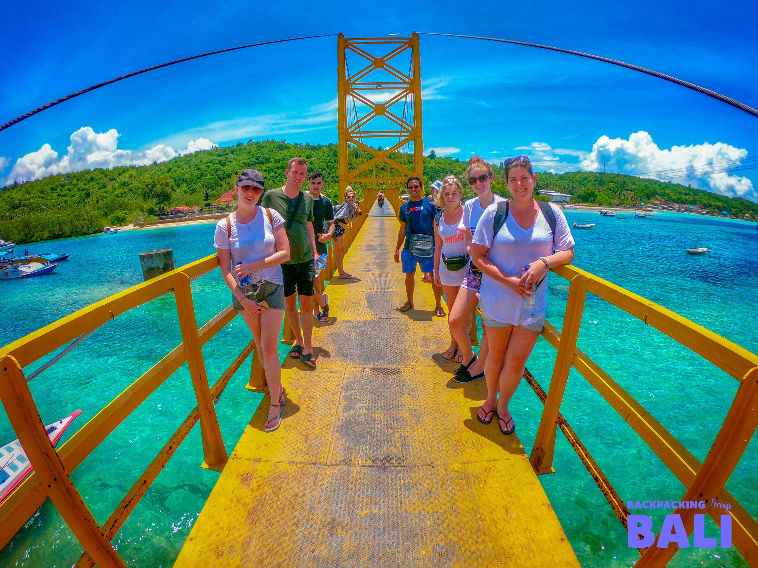 A group of happy backpackers pose on the iconic yellow bridge over turquoise water in Nusa Lembongan.
