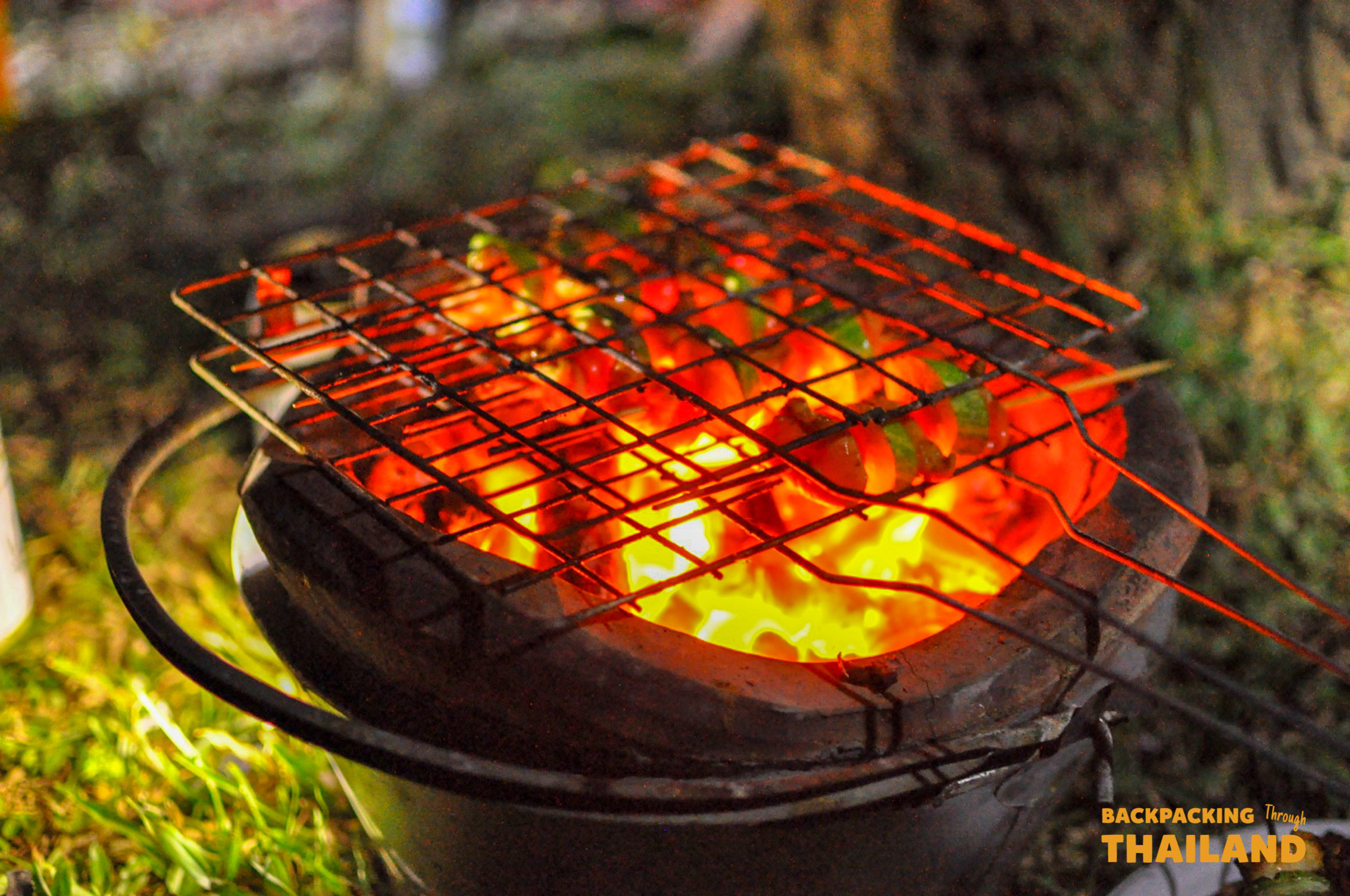 Close-up of colorful food skewers grilling over glowing red coals outdoors at night.