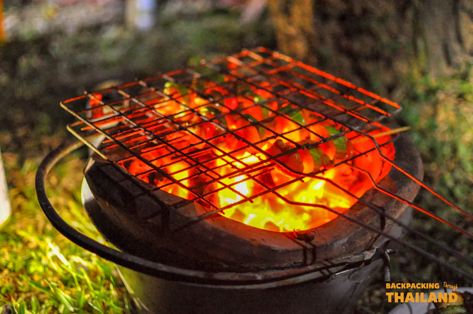 Close-up of colorful food skewers grilling over glowing red coals outdoors at night.